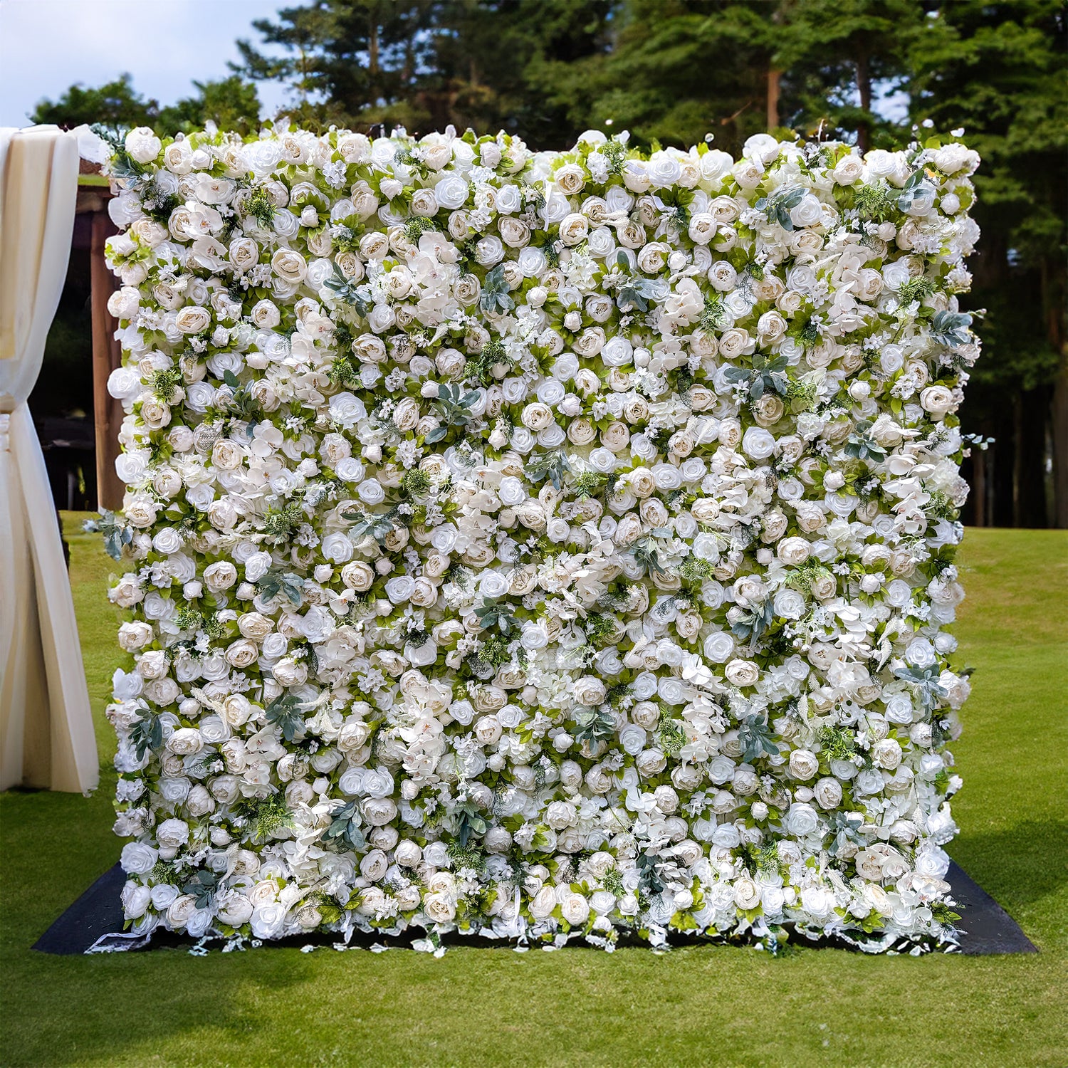 This image showcases a large, square - shaped flower wall. It is densely covered with white roses and lush green foliage. The flower wall stands on a well - maintained grassy area, with a backdrop of a forested landscape. The white roses are arranged in a way that creates a full and luxurious appearance, making the flower wall an ideal decoration for events such as weddings.