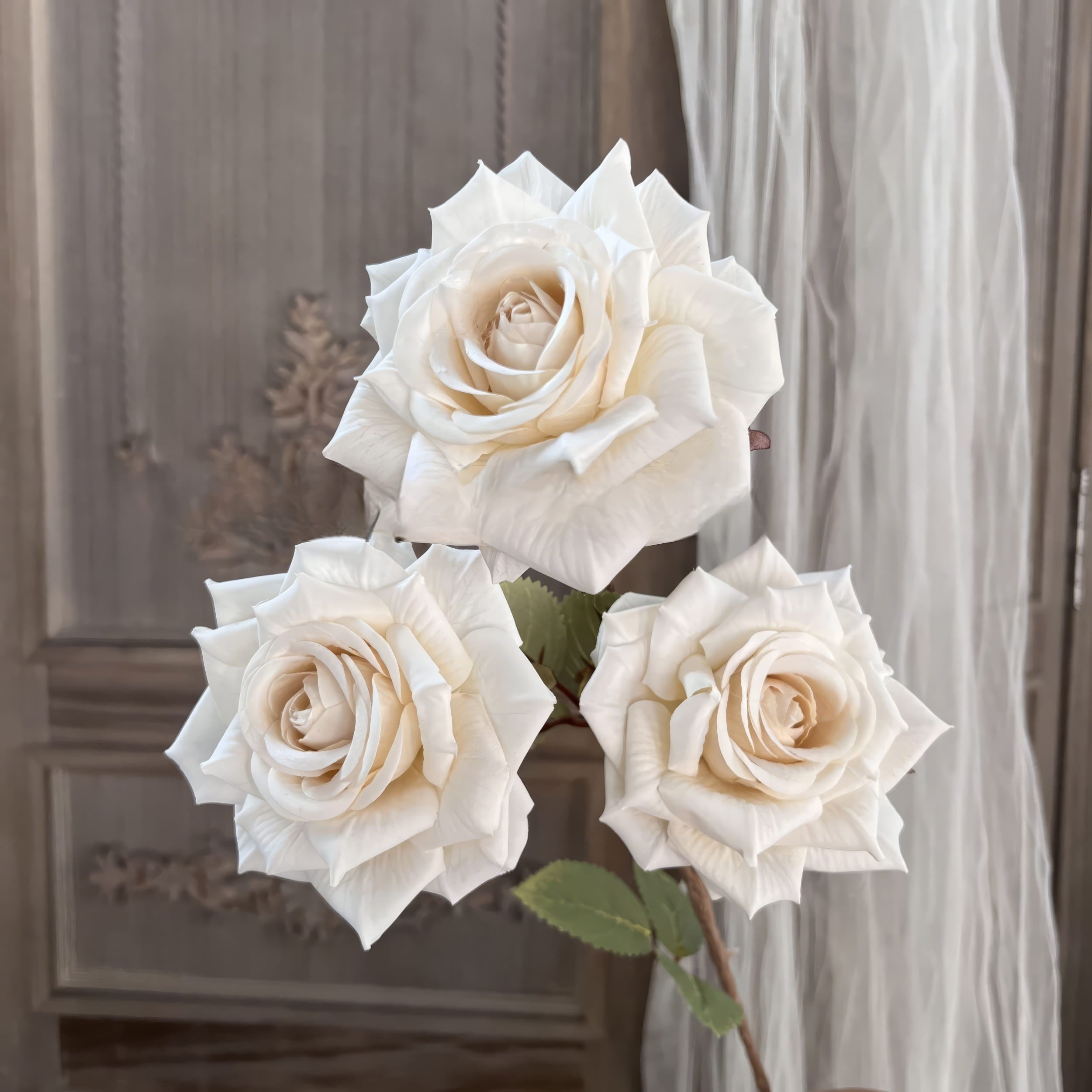 This image showcases three artificial roses in an elegant creamy white shade, with detailed petals and green leaves. The background features a soft, vintage-style wooden panel and sheer drapery for ambiance.