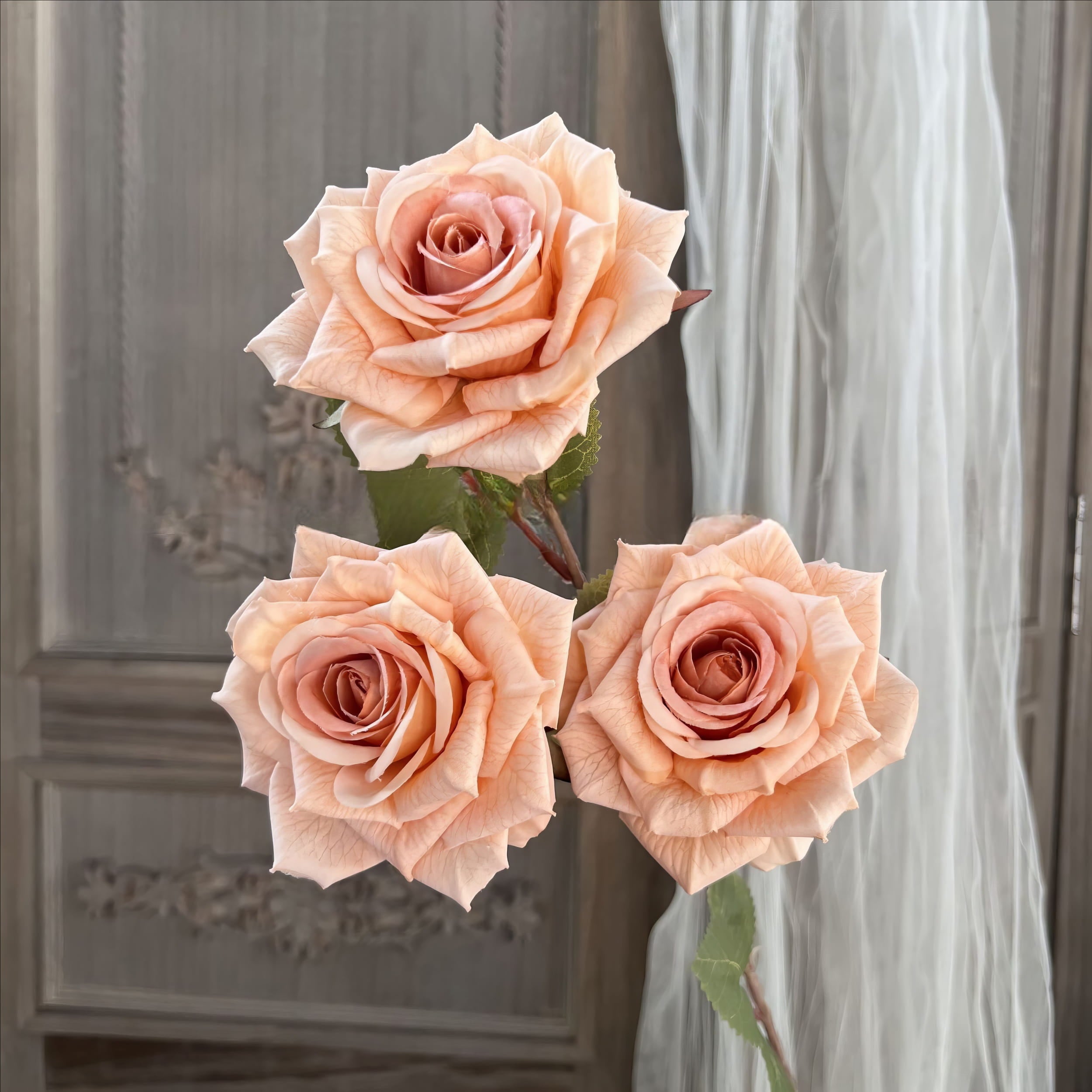 This image displays three lifelike artificial peach-colored roses with delicate petals and realistic textures. They are arranged on stems with green leaves, set against a soft, elegant background.