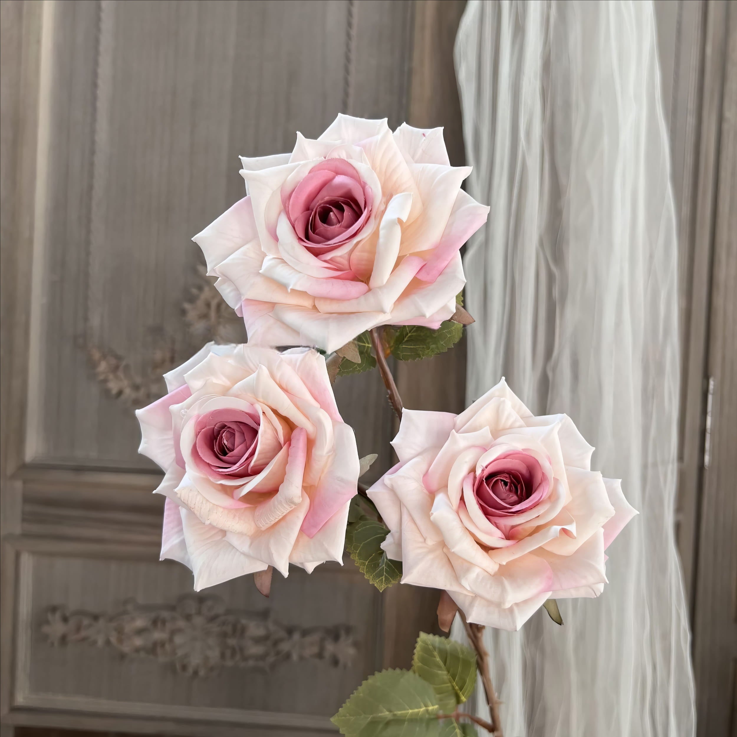 This image shows three beautifully crafted artificial roses with soft cream petals accented by subtle pink hues toward the center. The roses are realistic, with intricate petal textures and natural-looking green leaves, set against a sophisticated background.