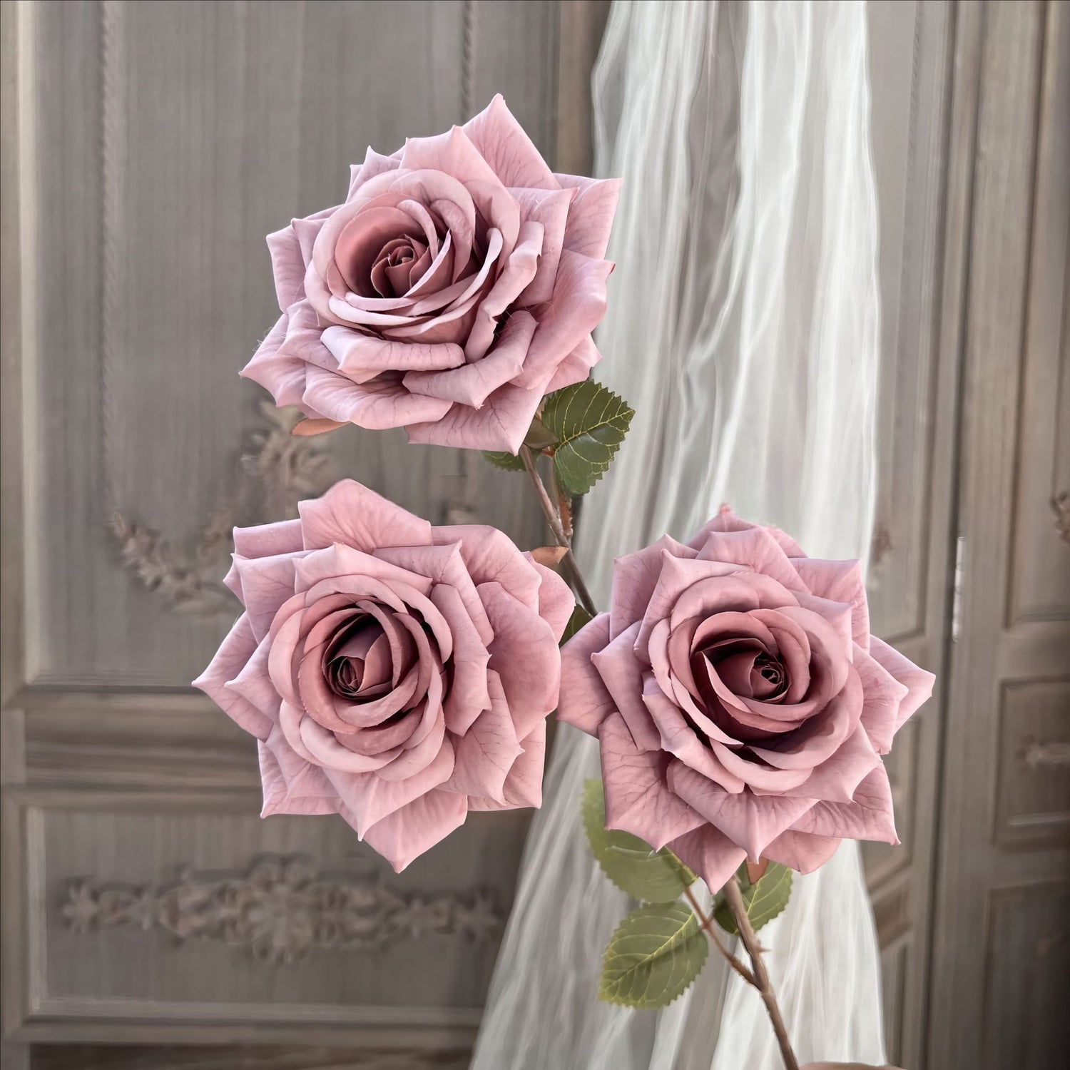 This image shows three elegant artificial roses in a dusty pink hue with realistic petals and green leaves. The background features a vintage wooden panel and sheer curtains, creating a soft, romantic ambiance.