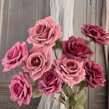 The image shows a bouquet of lifelike roses in varying shades of pink and red, with detailed petals and green leaves. The flowers are arranged against a vintage wooden background and sheer fabric drapery.