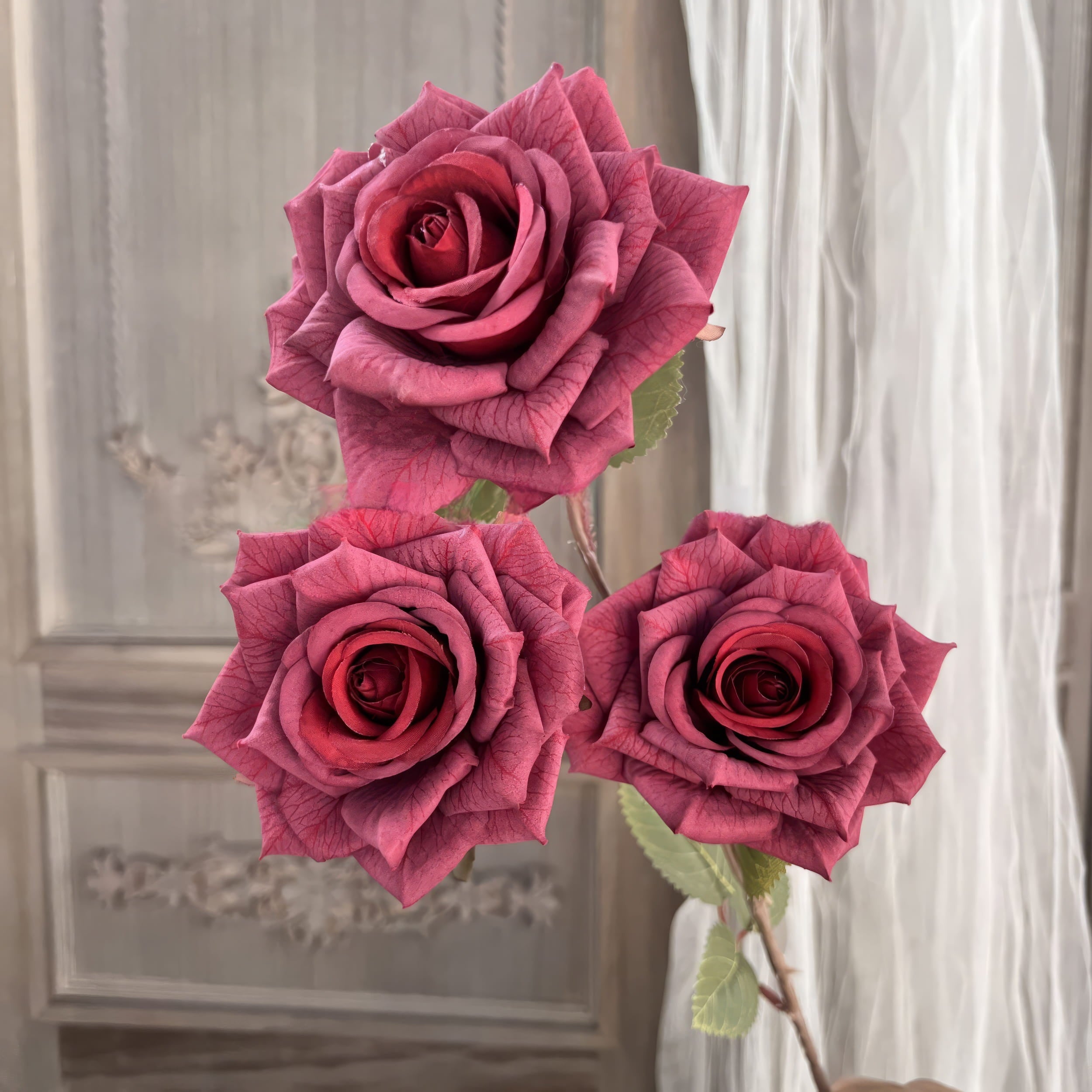 The image shows three deep red artificial roses with intricate, lifelike petals and green leaves. The background features a vintage wooden panel and sheer curtains, adding an elegant and romantic feel.