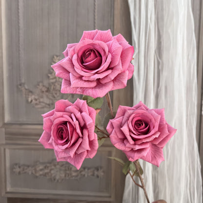 The image showcases three pink artificial roses with lifelike, textured petals and green leaves. The background features a vintage wooden panel and flowing sheer curtains, creating an elegant and delicate ambiance.