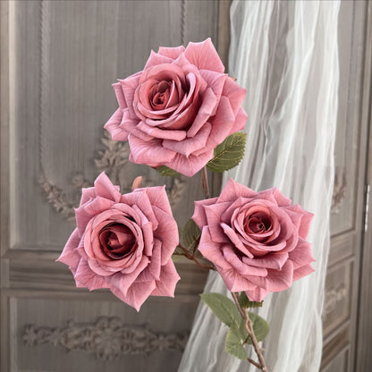 The image depicts three pink artificial roses with realistic, textured petals and detailed green leaves. They are set against a vintage wooden panel background, accented by sheer flowing curtains.