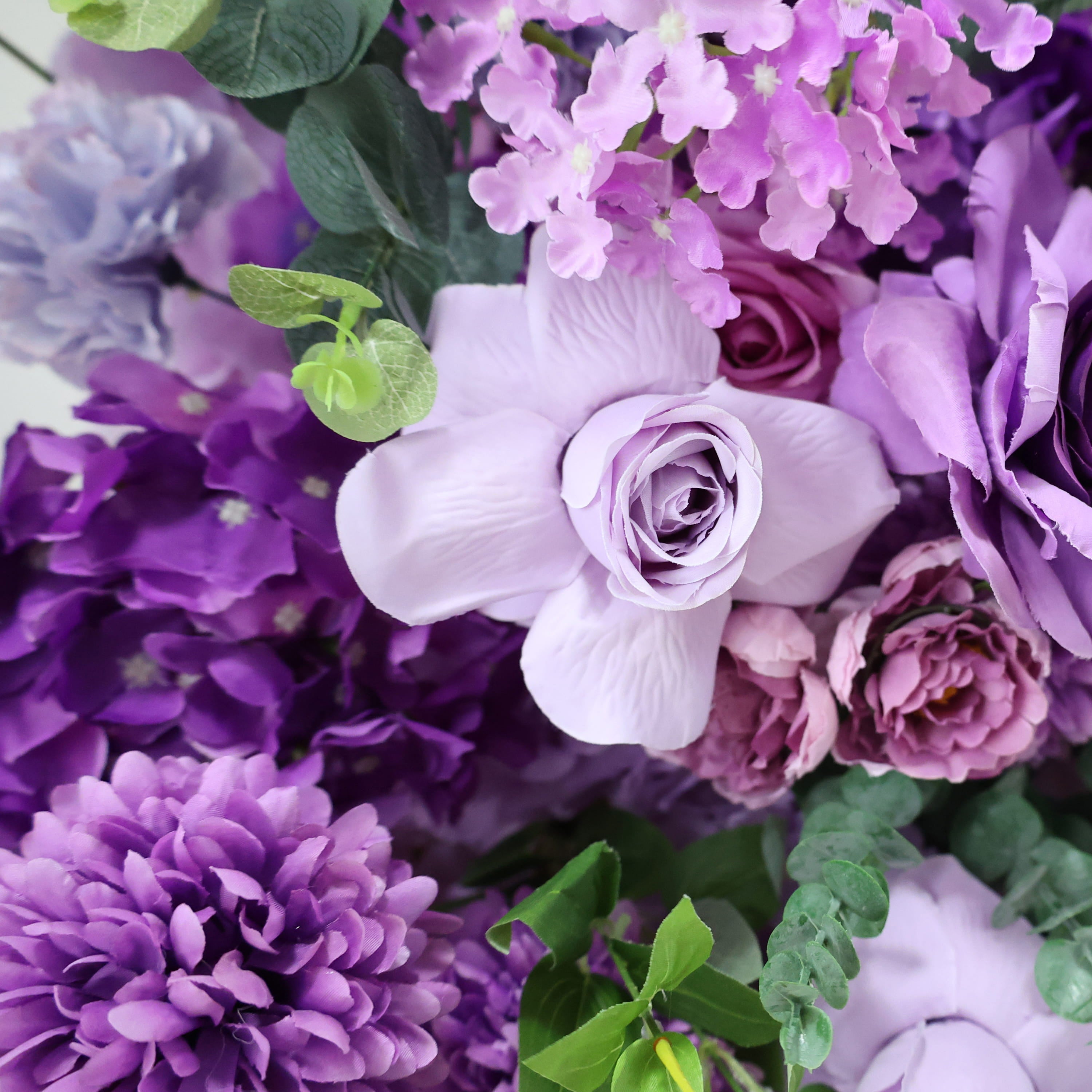 The ValarFlowers bouquet features a variety of flowers. It includes light purple roses, vibrant purple hydrangeas, and delicate lilac blooms, creating a rich and colorful arrangement.