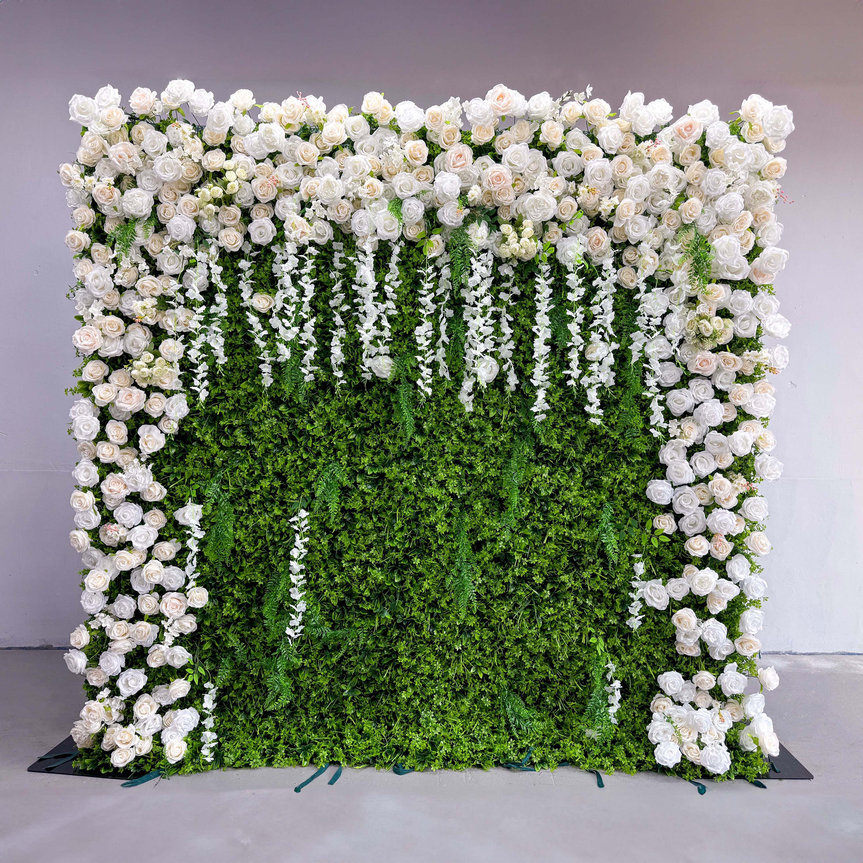 This flower wall has a green leafy background with a profusion of white roses. The roses are clustered together, especially at the top and sides. Hanging white floral strands add a dynamic element, contributing to the wall&