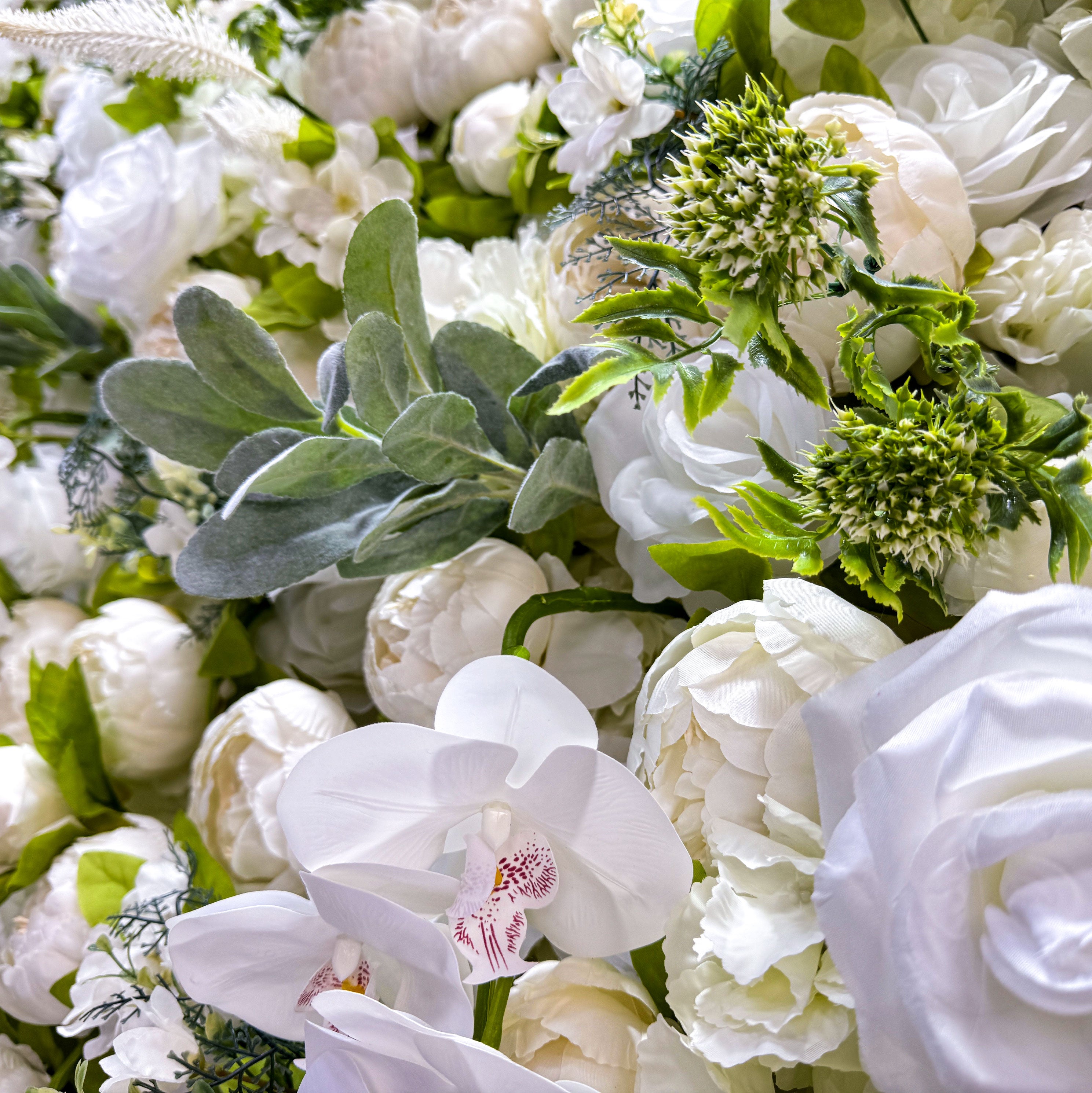 This close - up image showcases a variety of white and off - white flowers on a flower wall. The flowers are accompanied by green leaves, creating a lush and vibrant display. The close - up view highlights the intricate details of the petals, including the soft folds and natural - looking arrangements. The different types of flowers, such as roses and possibly peonies, add to the richness of the display.
