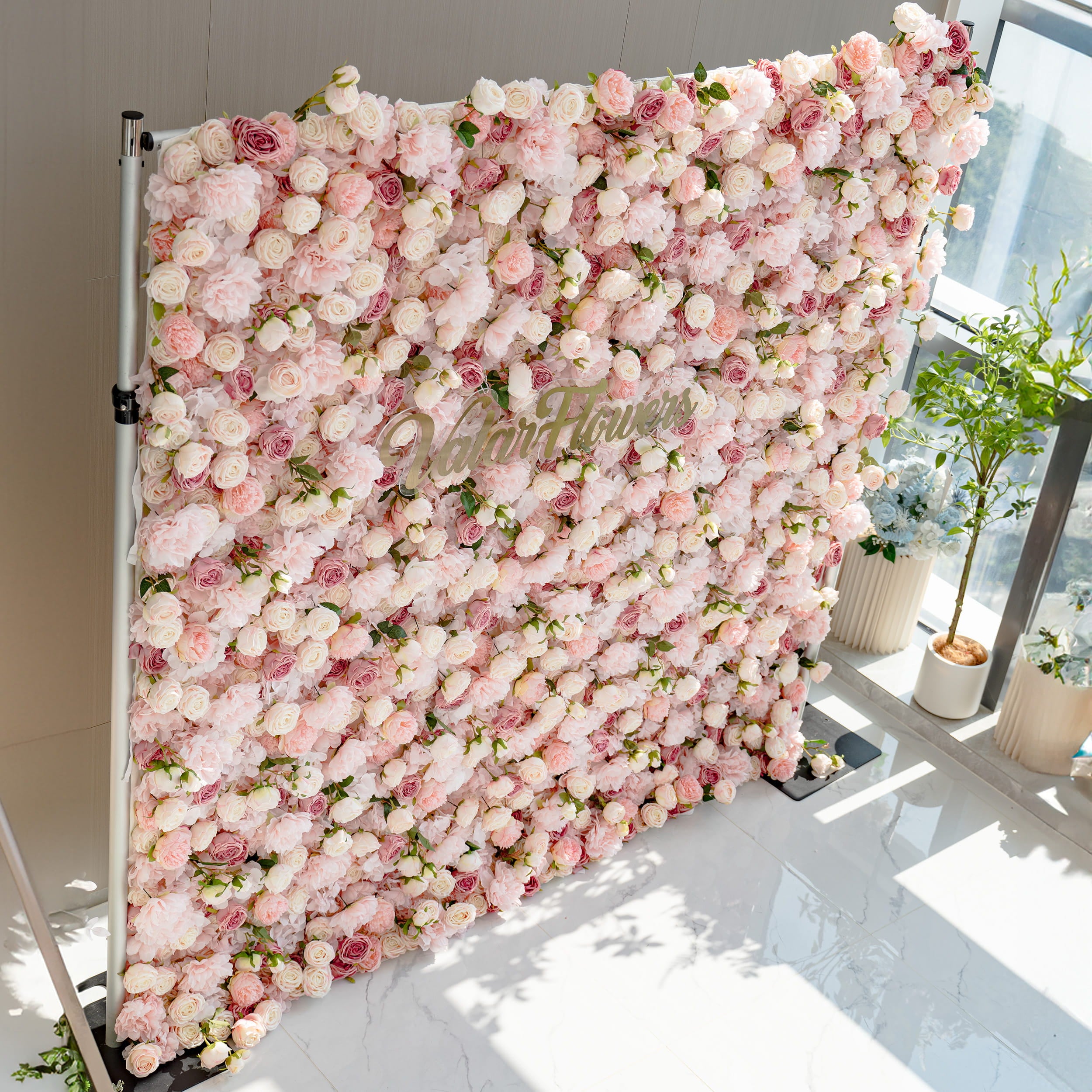 A stunning flower wall filled with a variety of pink and white roses. The wall is labeled &quot;ValarFlowers&quot; and is set against a modern interior with potted plants nearby.