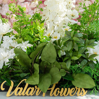 The floral arrangement features a variety of white flowers, including lilies and orchids, surrounded by lush green foliage. It is ideal for weddings, creating a romantic and elegant atmosphere. The sign &quot;Valar Flowers&quot; adds a charming touch.