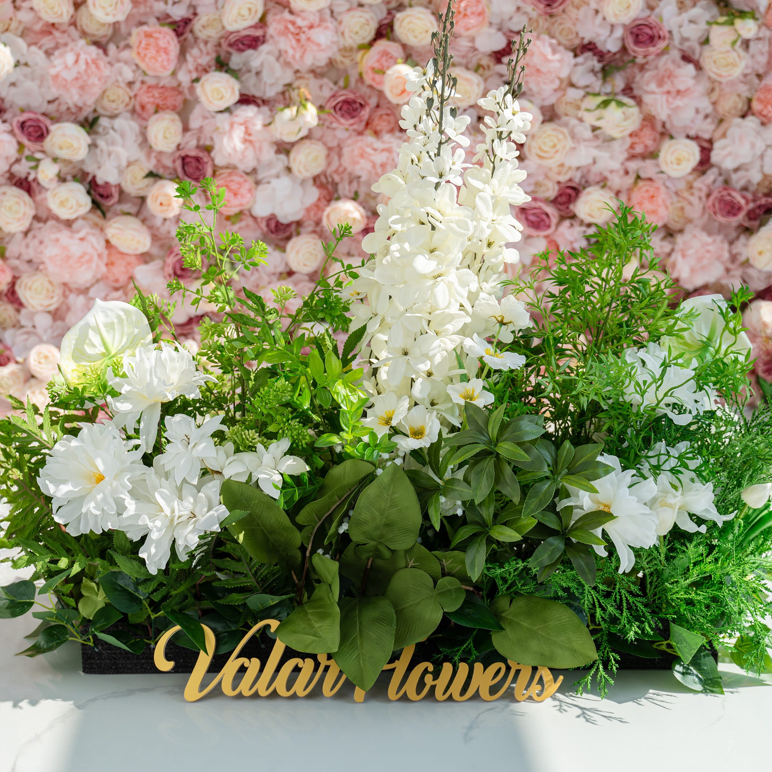 The floral arrangement features a variety of flowers, including white lilies, hydrangeas, and orchids, complemented by lush green foliage. It is ideal for weddings, creating a romantic and elegant atmosphere. The sign &quot;Valar Flowers&quot; adds a charming touch.