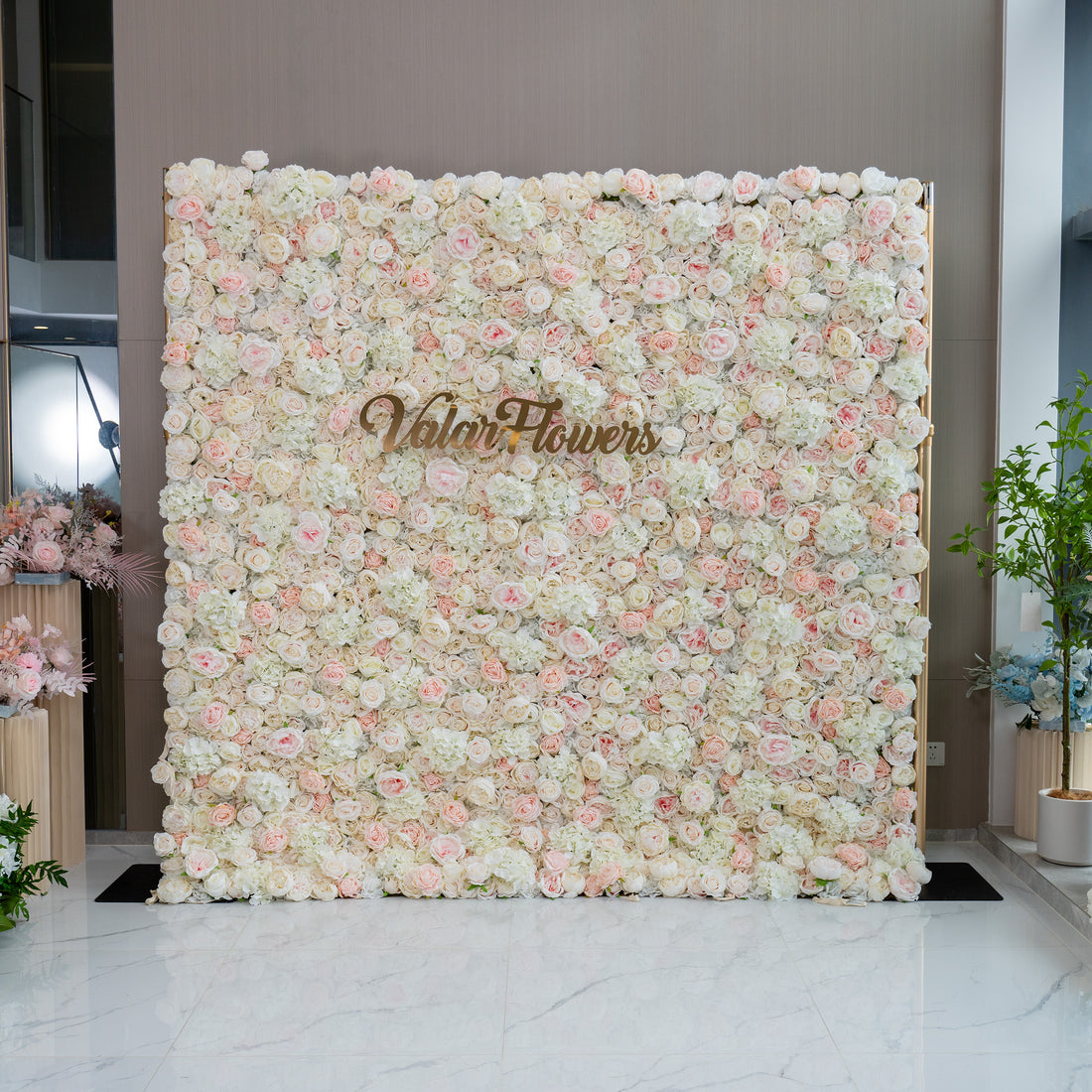 This image showcases a floral wall adorned with a dense arrangement of light pink and white flowers. The wall features the brand name &quot;ValarFlowers&quot; prominently displayed in the center. The overall ambiance is soft, romantic, and inviting, making it ideal for events such as weddings, bridal showers, or romantic celebrations.