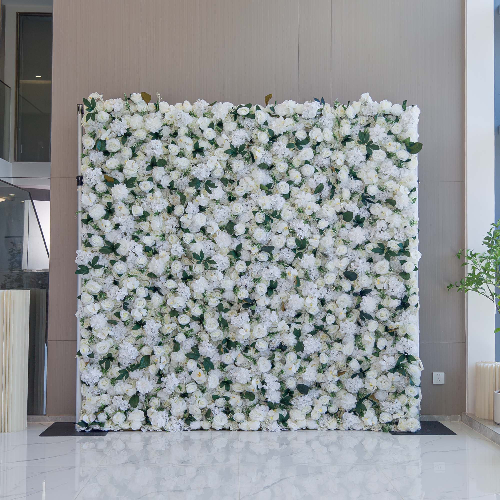 A beautifully arranged flower wall decor featuring a dense display of white roses and hydrangeas, interspersed with green leaves. The backdrop is complemented by a sleek, modern interior, enhancing its elegance. Ideal for events or as a decorative accent.