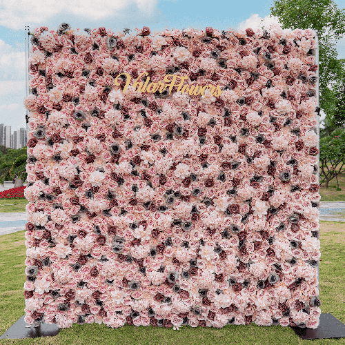 This flower wall is densely packed with pink-hued roses, each accentuated with dark, intricate details in their centers. The uniform design offers a sumptuous visual texture. A golden &quot;Valar Flowers&quot; sign crowns the top, contrasting the blooms. Positioned outdoors, it stands as a vivid focal point against a city skyline backdrop.