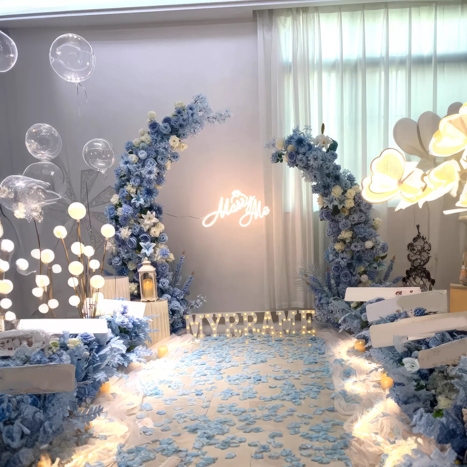 A dreamy blue floral arch with a &quot;Marry Me&quot; sign, surrounded by glowing orbs and delicate balloons. The aisle is sprinkled with petals, creating a whimsical and romantic ambiance for a memorable proposal.