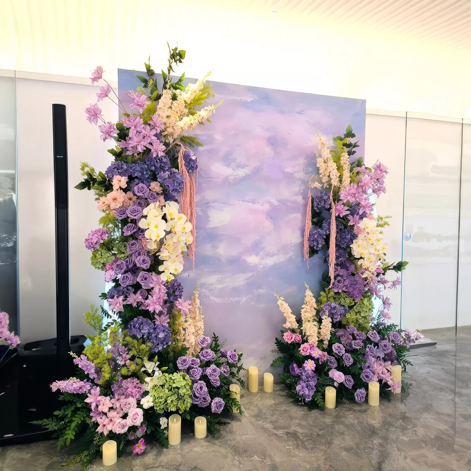 A vibrant floral arch featuring purple and pink blooms against a pastel backdrop. Accented with candles, this enchanting setup offers a serene and romantic atmosphere, perfect for weddings or special events.