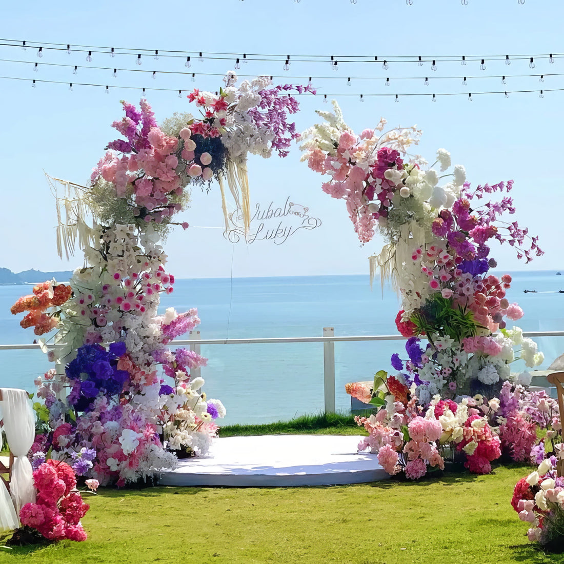 A stunning floral moon gate arch stands against a serene ocean backdrop. Adorned with vibrant pink, purple, and white blooms, it creates a romantic setting perfect for weddings or special occasions, enhanced by string lights overhead.