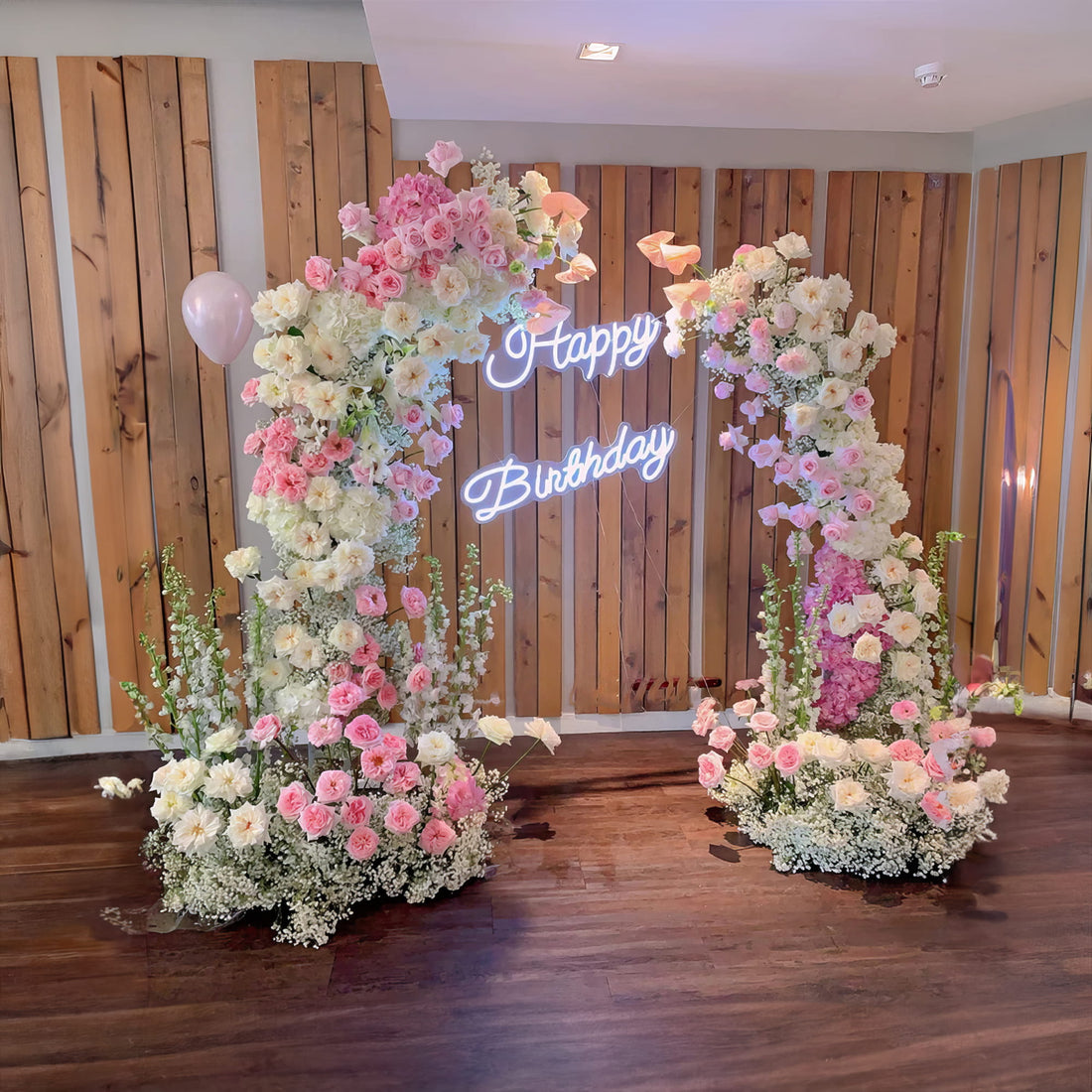 A charming floral arch with pink and white roses frames a neon &quot;Happy Birthday&quot; sign. Set against a wooden backdrop, it creates a festive and elegant ambiance, perfect for a birthday celebration.