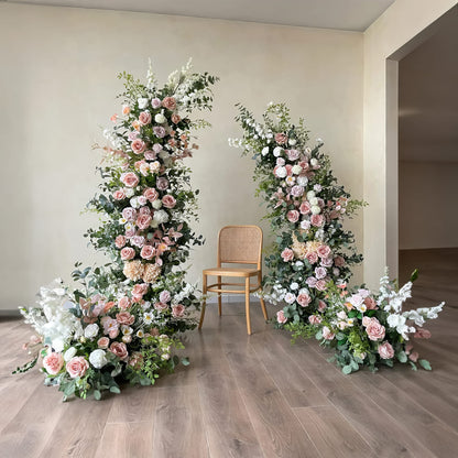 Elegant floral arrangements featuring blush pink and white roses with lush greenery frame a simple wooden chair. Set against a neutral backdrop, this setup is perfect for creating a serene and sophisticated ambiance in any event space.