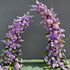 Magnificent artificial floral arch for weddings, ceremonies, and special events. Features vibrant purple and lavender flowers (roses, peonies, hydrangeas, lilacs) with lush greenery. Arranged in a cascading effect on a durable frame. Ideal for indoor/outdoor use. Luxurious, romantic design.