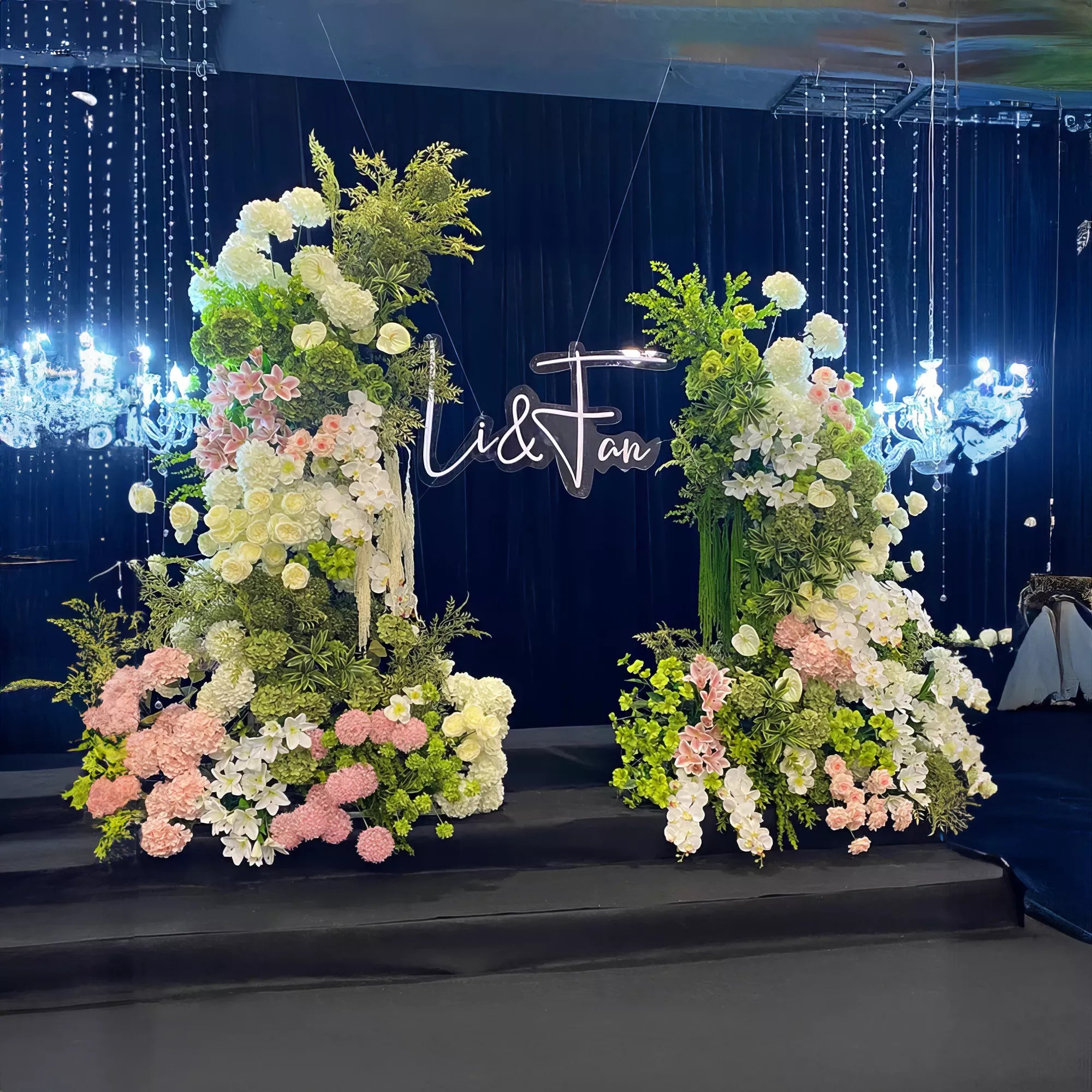 A stunning floral wedding arch adorned with lush greenery and blooms in white, pink, and green hues. Elegant chandeliers and cascading lights enhance the romantic ambiance, while a personalized sign adds a unique touch to this enchanting setup.