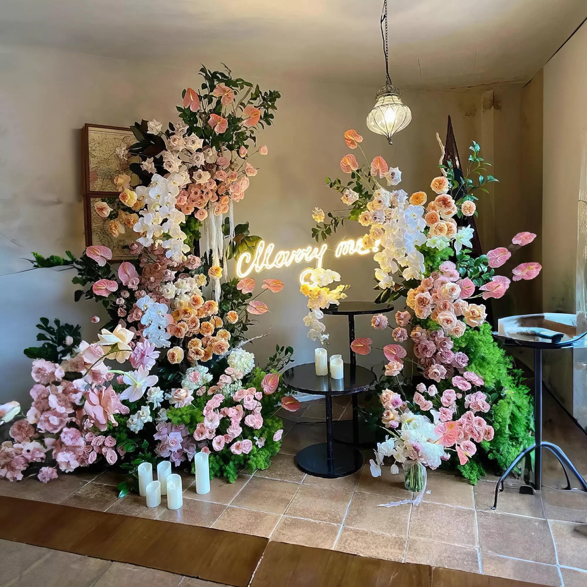 A charming floral arch featuring pink and white blooms with lush greenery, accented by a glowing &quot;Marry Me&quot; sign. Soft candlelight and a vintage hanging lamp create a cozy, romantic atmosphere, perfect for proposals and intimate celebrations.