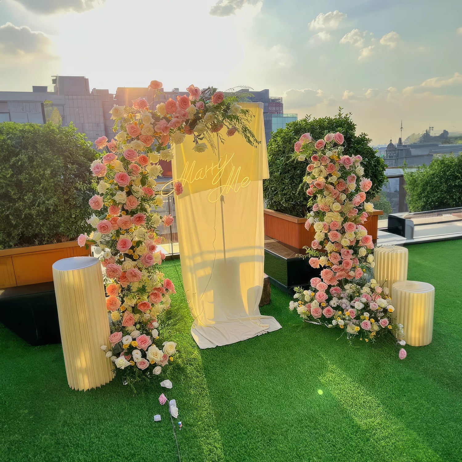 A beautiful floral arch adorned with pink and white roses, set on a rooftop with a &quot;Marry Me&quot; sign. The sunlit scene with lush greenery and elegant lighting creates a perfect romantic setting for proposals and special moments.