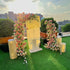 A beautiful floral arch adorned with pink and white roses, set on a rooftop with a "Marry Me" sign. The sunlit scene with lush greenery and elegant lighting creates a perfect romantic setting for proposals and special moments.