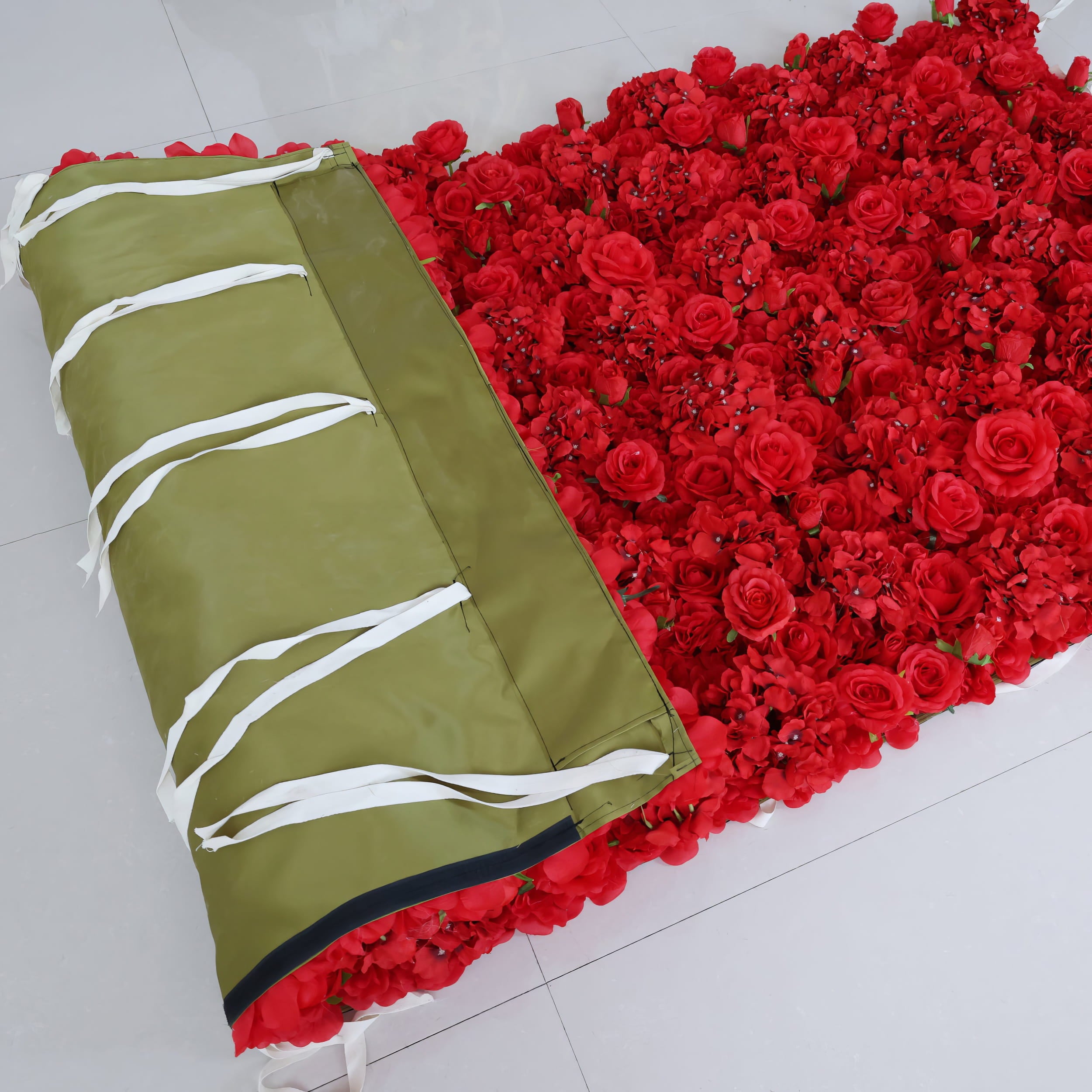 A partially rolled ValarFlowers red flower wall, showcasing vibrant silk blooms on the front and a durable olive-green fabric backing with white tie straps for secure attachment at events or venues.