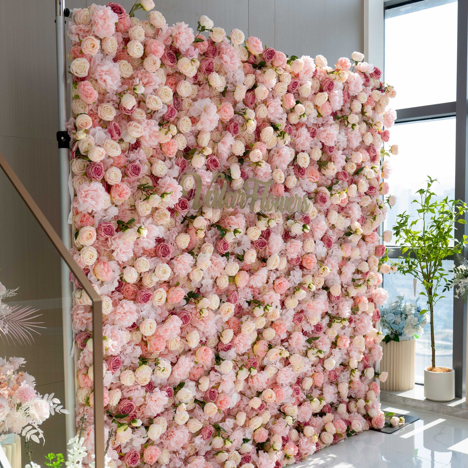 A large, square flower wall filled with pink and white roses and peonies. It has the text &quot;Valar Flowers&quot; on it. Surrounding plants add to the floral ambiance.