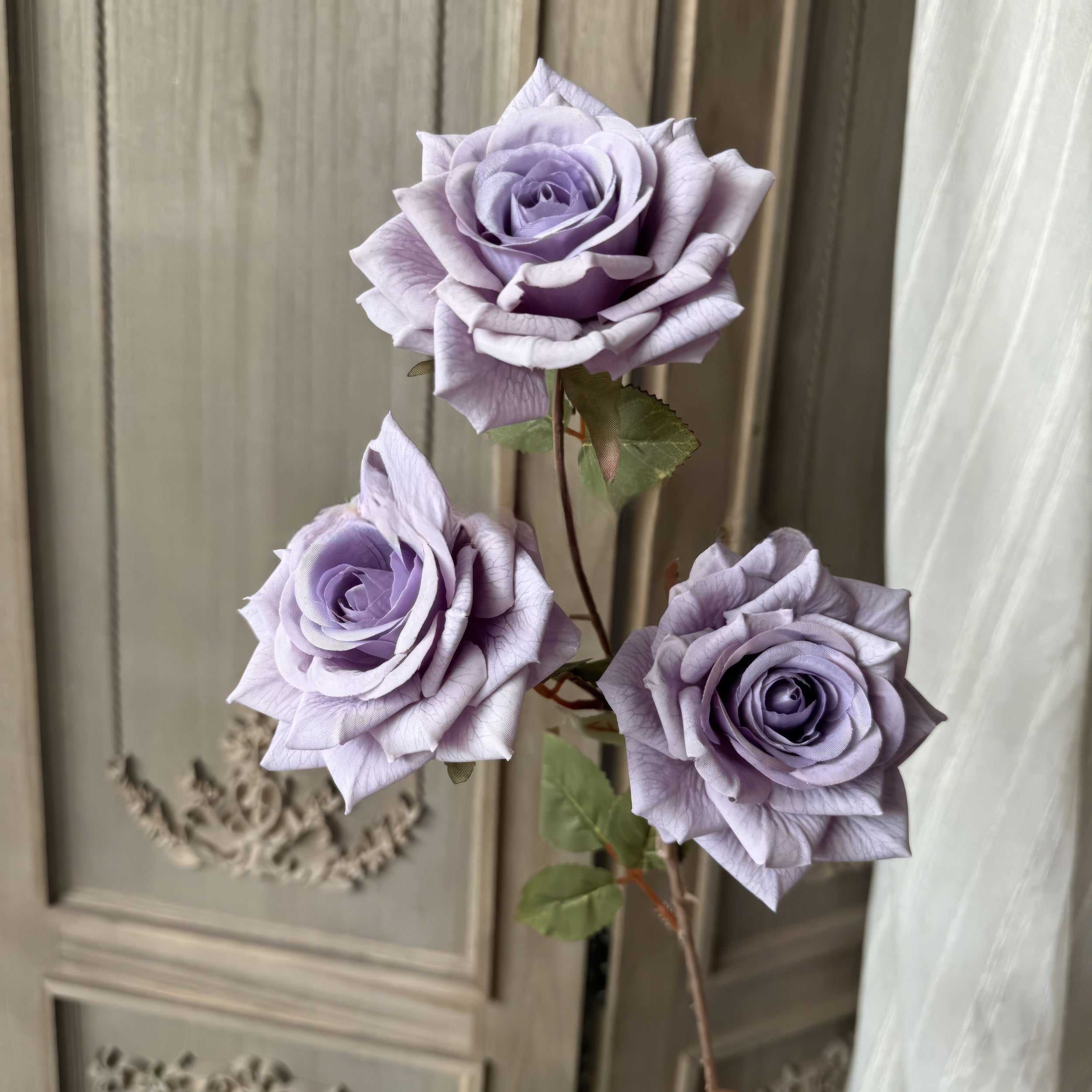 This image features three lavender roses in full bloom with delicately detailed petals. The roses are accompanied by green leaves, set against a beige textured background with ornate decorative accents.