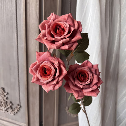 This image showcases three elegant deep pink roses in full bloom, with intricate petal textures. They are accompanied by green leaves, set against a neutral background with soft fabric details.