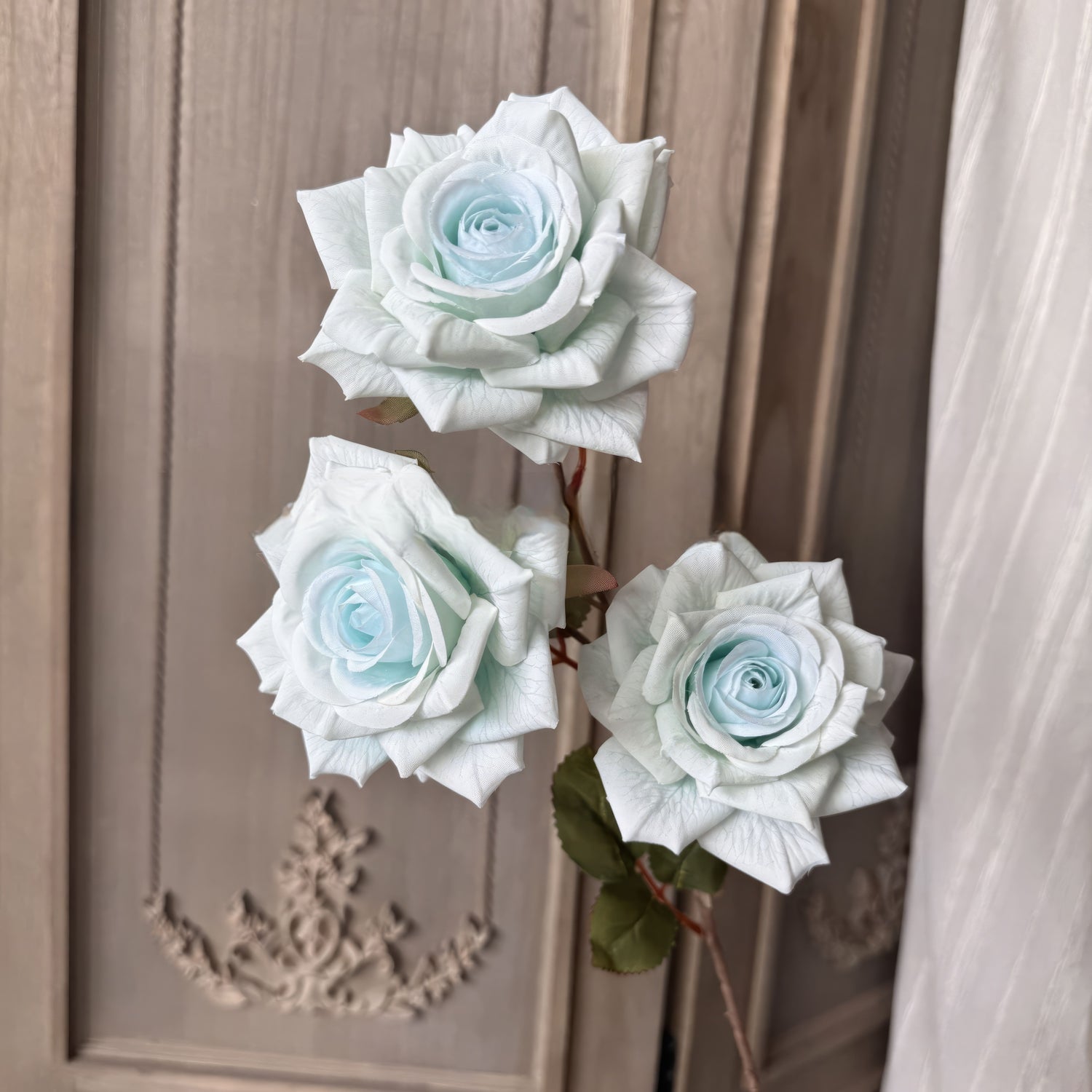 This image showcases three pale blue roses with intricate, delicate petals arranged in full bloom. The roses are accompanied by green leaves, set against a textured beige background with ornate detailing.