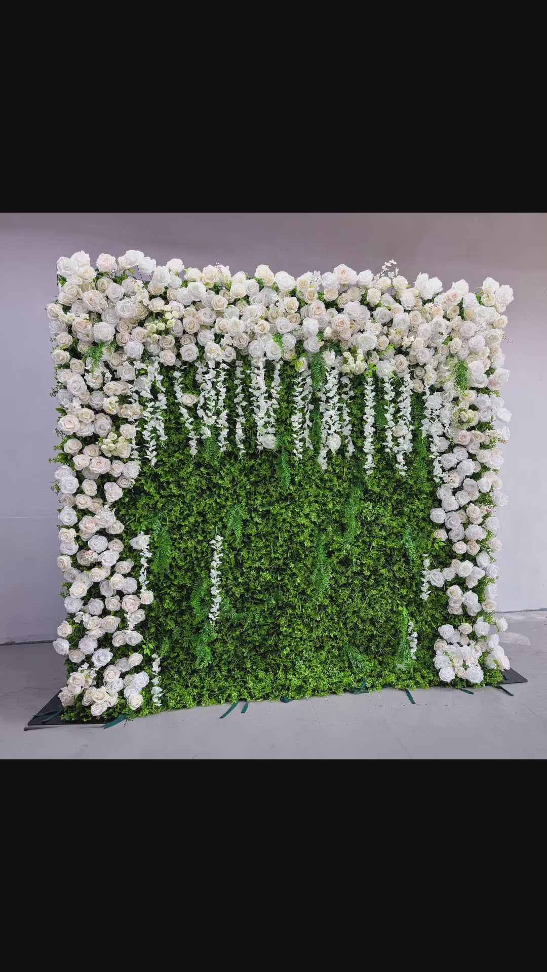 The flower wall features a green foliage base with numerous white roses. The roses are arranged to cover the top and sides, while white floral strands hang down the front. The combination of green and white creates a fresh and elegant look.